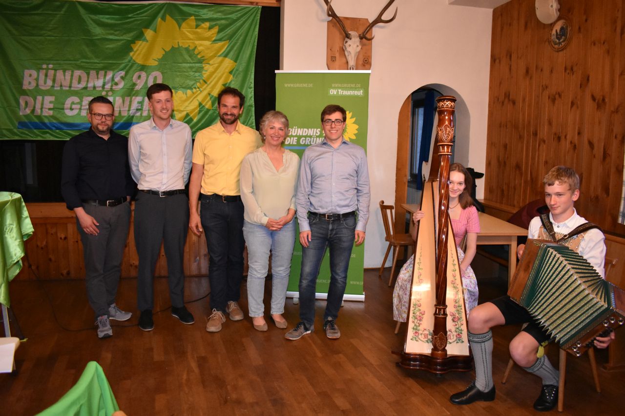 In einem Raum stehen fünf Menschen in einer Reihe. Rechts daneben sitzen eine Harfenspielerin und ein Junge mit Akkordeon. Im Hintergrund sieht man Fahnen der Partei Bündnis 90, die Grünen.