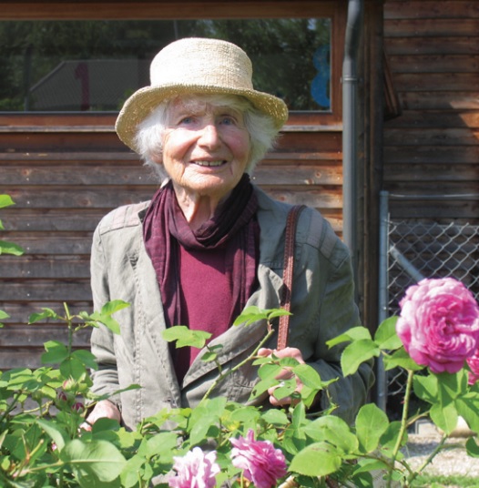 Vor einem Holzhaus steht eine ältere Dame mit Hut, vorne blühen rosa Rosen.