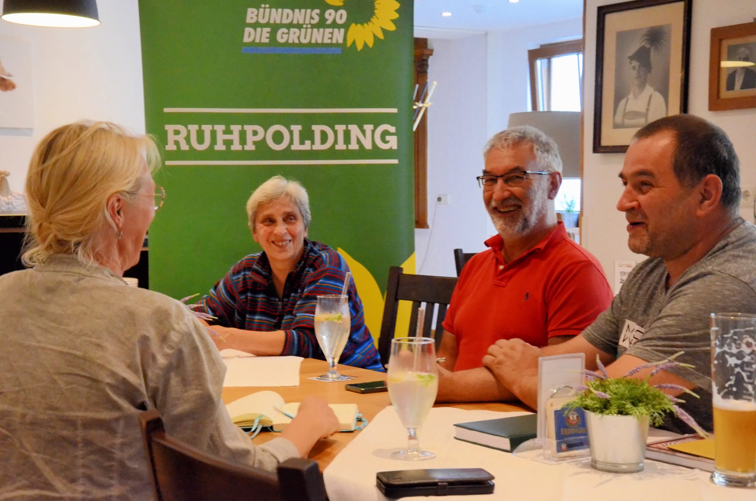 Um einen Wirtshaustisch sitzen vier Menschen in gelöster Stimmung. Im hHintergrund ein Banner der Grünen OV Ruhpolding.