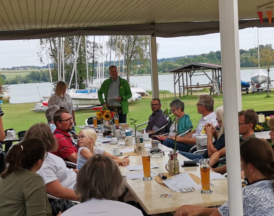 Zwei Menschen stehen vor einer Gruppe, die gemütlich an einer langen Tafel sitzen. Im Hintergrund der Waginger See und Segelboote