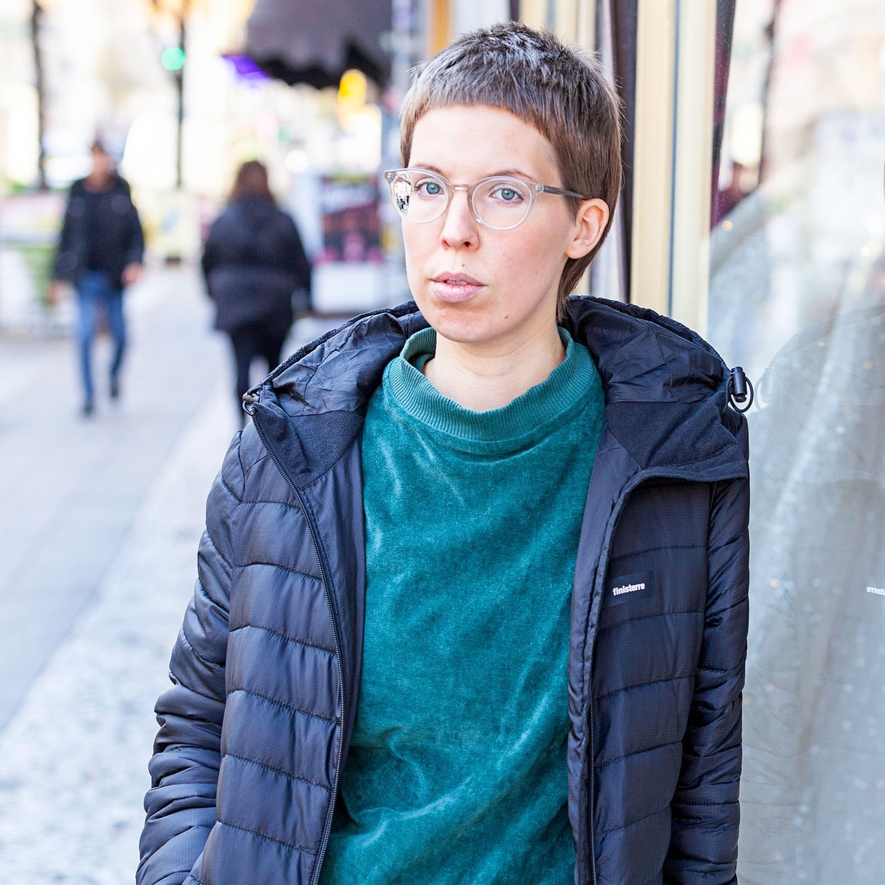 An ein Schaufenster gelehnt steht eine Frau mit kurzen Haaren und Brille. Sie spiegelt sich im Schaufensterglas.